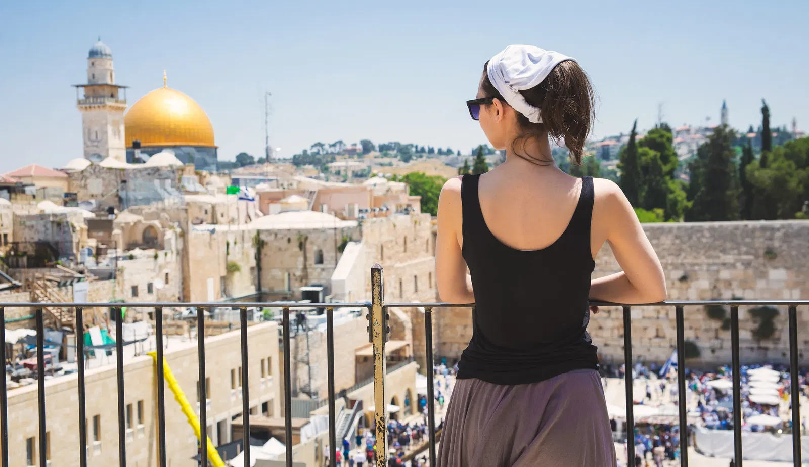 A woman with a hat looking at the old city of Jerusalem from a balcony.